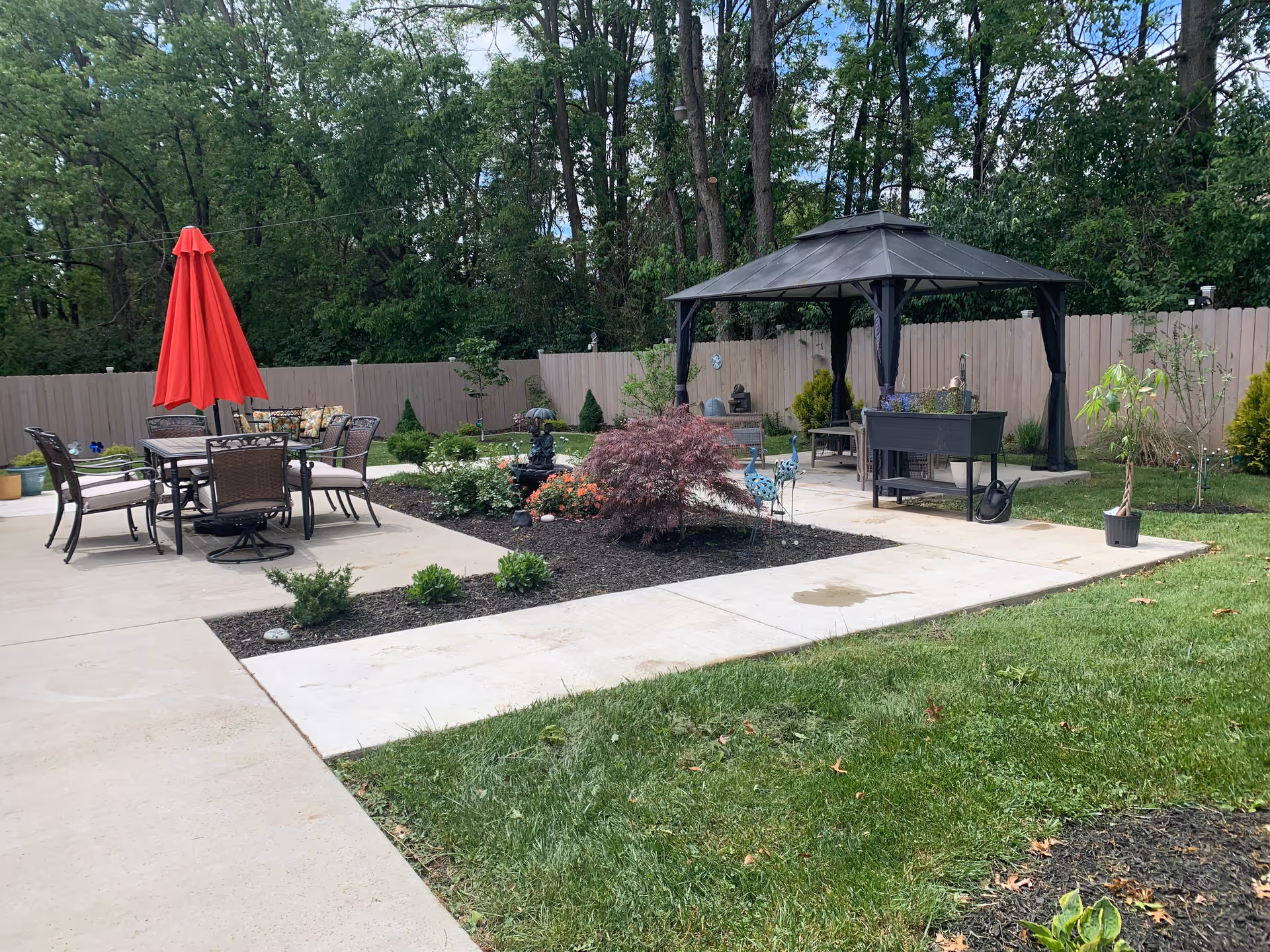 Outdoor patio area with a dining table and chairs under a red umbrella on the left, a garden bed with various plants and a small fountain in the center, and a gazebo with seating and a planter on the right. The area is surrounded by a wooden fence and trees in the background.