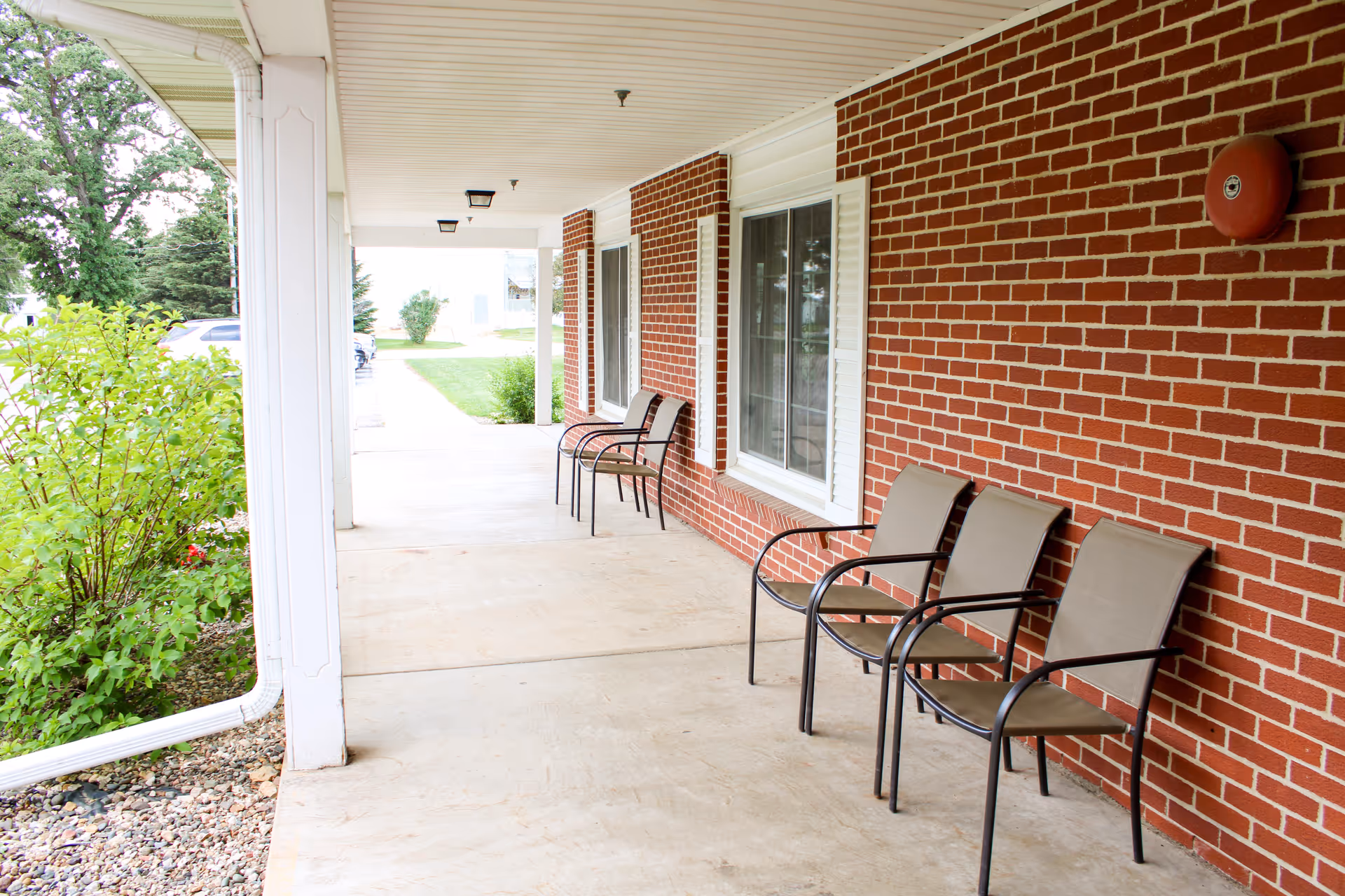 Covered brick porch with several outdoor chairs along a concrete walkway and greenery to the side.