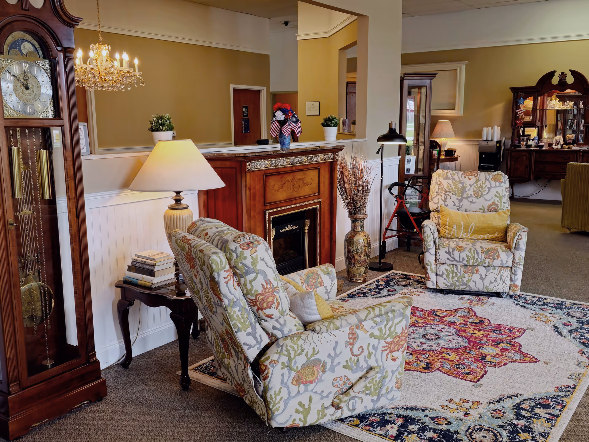 A cozy living room area in Crown Assisted Living featuring two floral-patterned armchairs with yellow pillows, a colorful area rug with intricate designs, a wooden grandfather clock, a side table with stacked books and a lamp, a decorative fireplace with a vase of flowers on top, and a floor lamp next to a red walker. The room has warm beige walls and carpeted flooring.