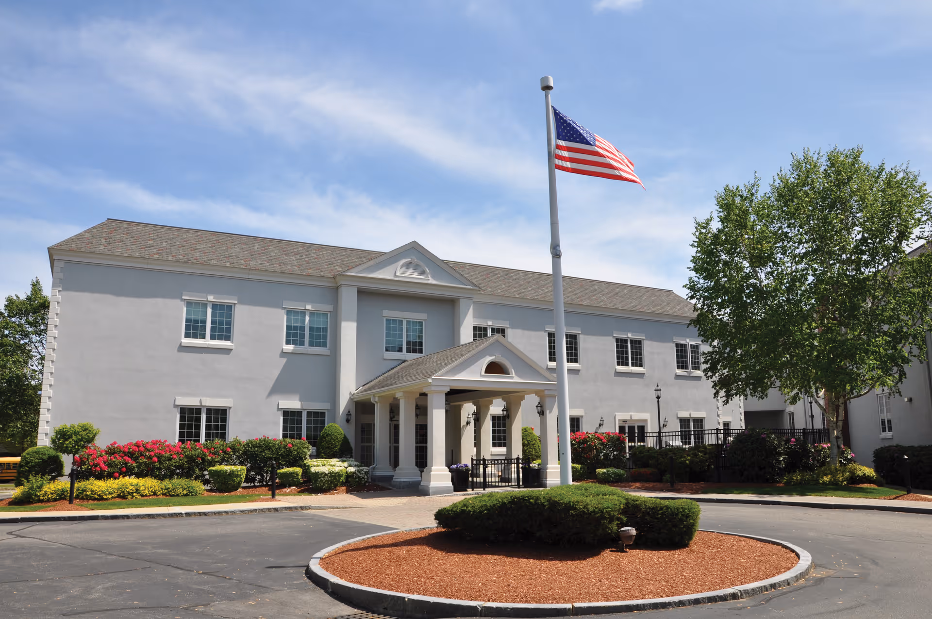 Exterior view of a two-story healthcare center building with a covered entrance supported by columns. There is a circular driveway with a landscaped island in the center featuring bushes and a flagpole flying the American flag. The building is surrounded by well-maintained shrubs, flowers, and trees under a partly cloudy blue sky.