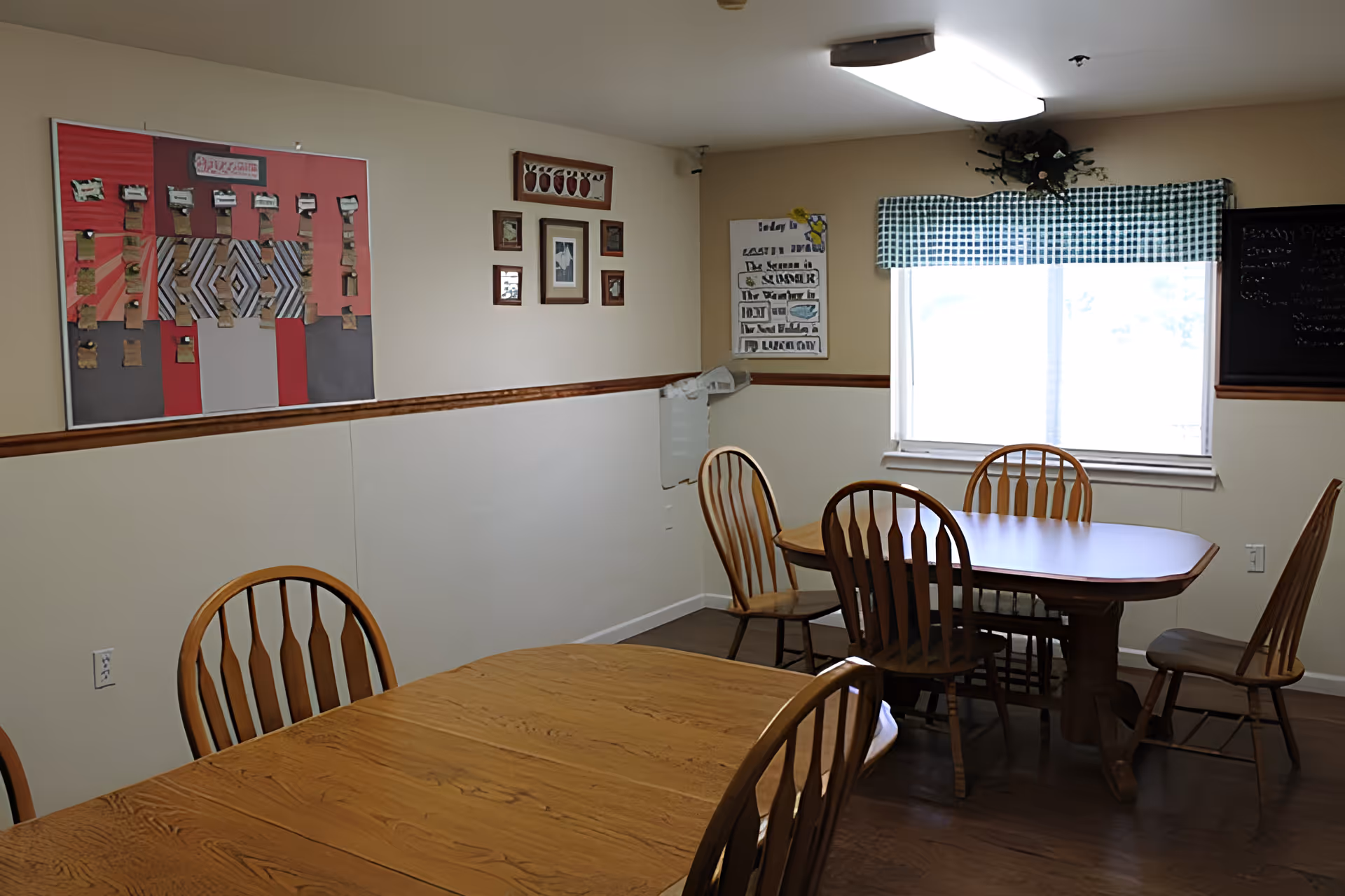 A dining room with two wooden tables and several wooden chairs. The walls are beige with a wooden trim. There is a window with a green and white checkered valance letting in natural light. Various framed pictures and a bulletin board with papers are displayed on the walls.