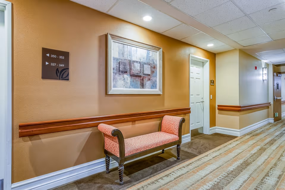 A well-lit hallway in a senior living facility with beige and light brown walls, a patterned carpet, a cushioned bench with wooden legs and armrests, a framed abstract painting on the wall, directional signs indicating room numbers, and a white door.