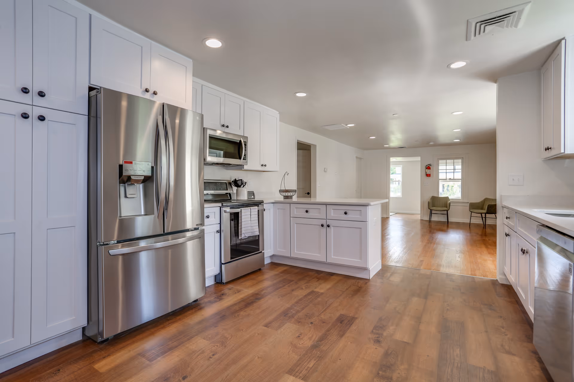 Open modern kitchen with stainless steel refrigerator and stove, white cabinetry and an island, leading into a bright living area with hardwood floors.