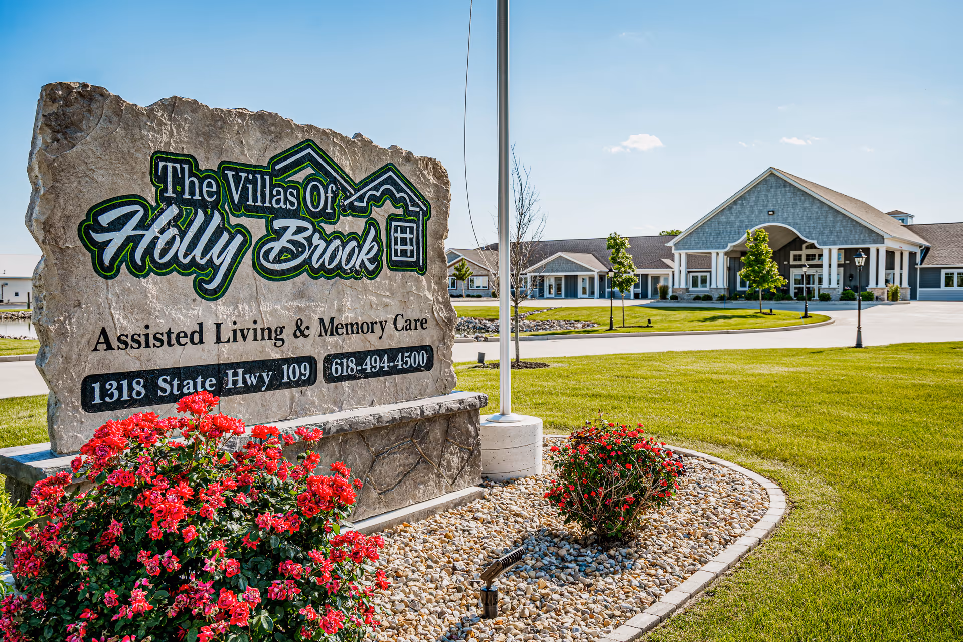 Large stone sign for The Villas of Holly Brook Assisted Living & Memory Care with address 1318 State Hwy 109 and phone number 618-494-4500, surrounded by red flowers and landscaping, with the facility building visible in the background under a clear blue sky.