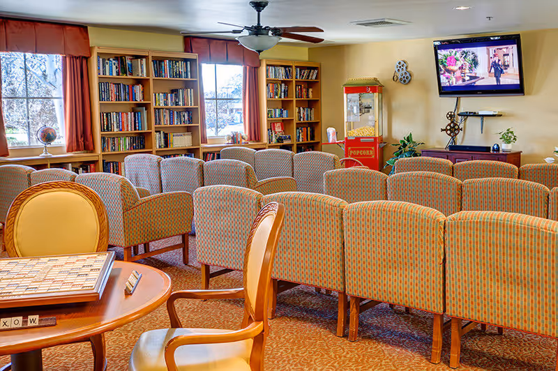 A cozy common room with rows of patterned chairs facing a wall-mounted television. The room has large windows with red curtains, bookshelves filled with books, a popcorn machine, and a round table with a Scrabble board game and chairs in the foreground.