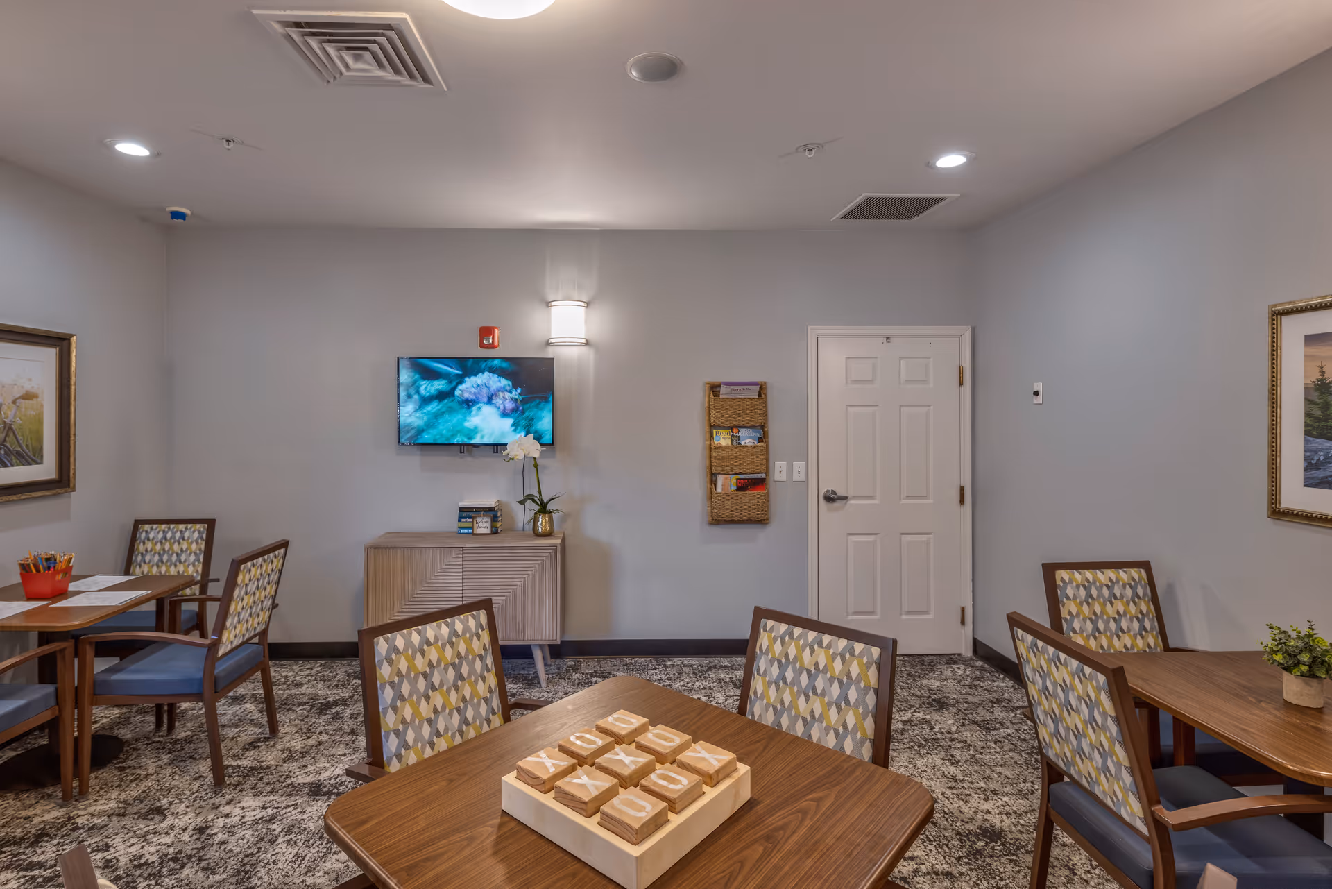 A well-lit interior room with multiple wooden tables and chairs featuring patterned upholstery. On one table, there is a wooden tic-tac-toe game. The walls are painted light gray and decorated with framed artwork. A flat-screen TV is mounted on the wall above a small cabinet, and a wall-mounted organizer holds brochures. The room has a carpeted floor and a closed white door.