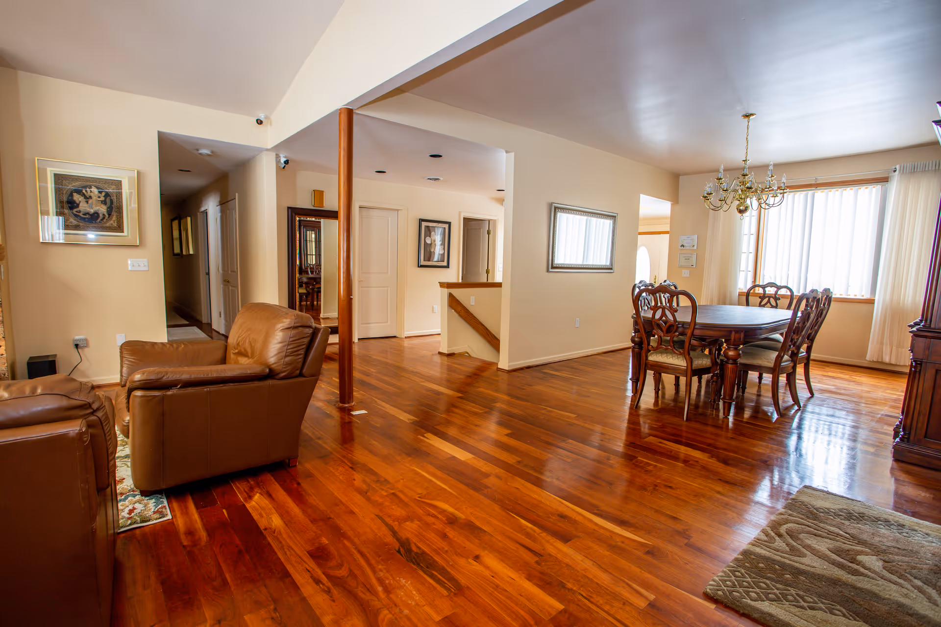 A spacious interior room with polished wooden floors featuring a dining area with a wooden table and six chairs under a chandelier near large windows with vertical blinds. To the left, there are two brown leather armchairs and a hallway leading to other rooms. The walls are painted beige and decorated with framed artwork and a mirror.