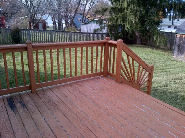 View of a wooden deck with a decorative railing overlooking a fenced backyard with grass, trees, and neighboring houses in the background.