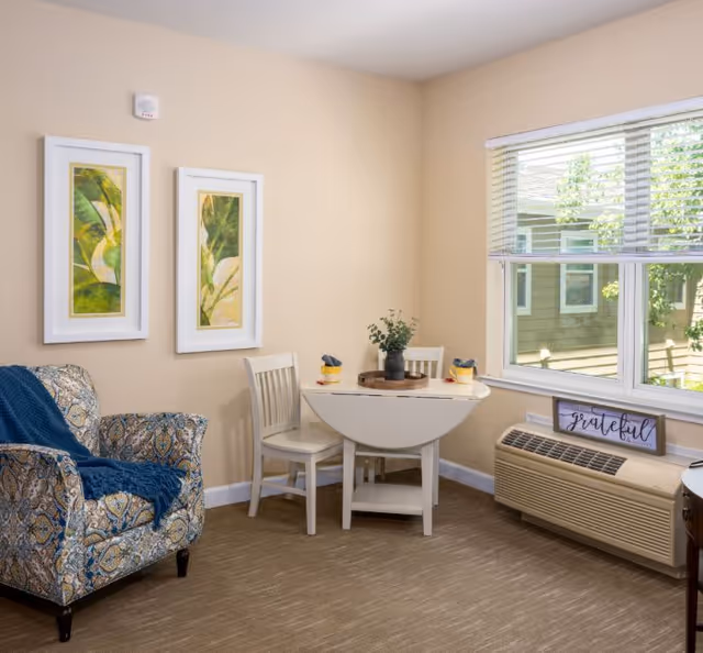 Cozy seating area with a patterned armchair, a small drop-leaf table and two chairs next to a window and wall art.