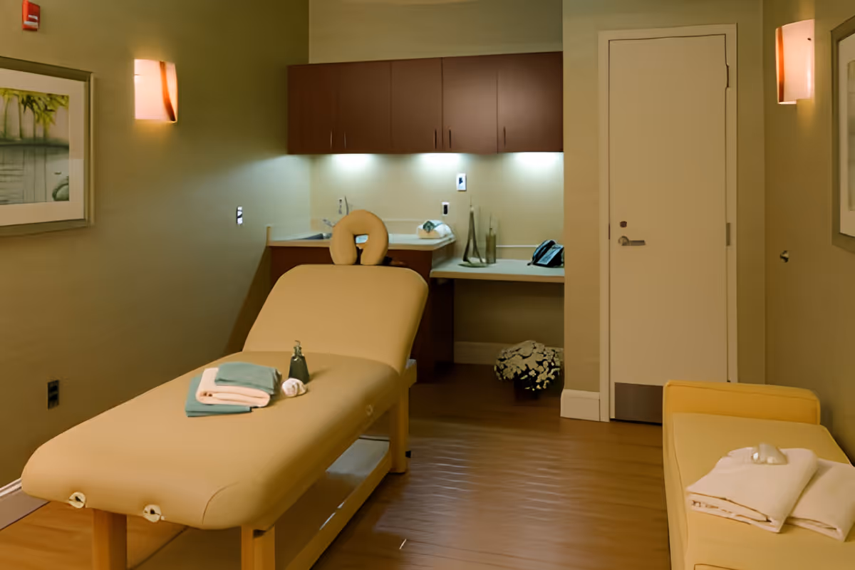 A serene massage or therapy room with a beige massage table in the center, topped with folded towels and a bottle of lotion. The room has wooden flooring, soft wall lighting, a bench with additional folded towels, and a countertop with cabinets and decorative items in the background.