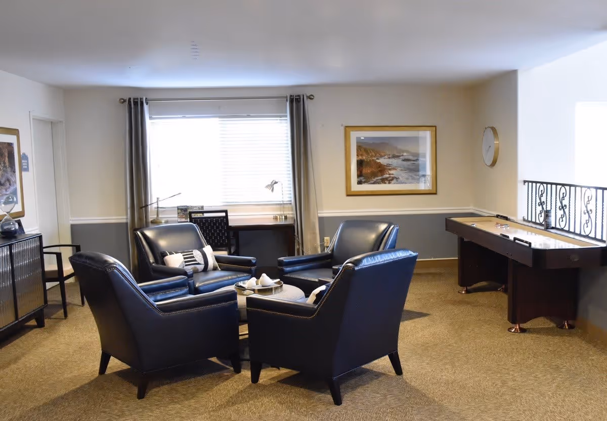 A cozy sitting area in a senior living facility with four dark leather armchairs arranged around a small round table. Behind the chairs is a window with closed blinds and curtains, a desk with a chair and a lamp. On the right side, there is a shuffleboard table next to a railing with decorative ironwork. The walls are painted in two tones with a framed landscape picture and a clock hanging on them.