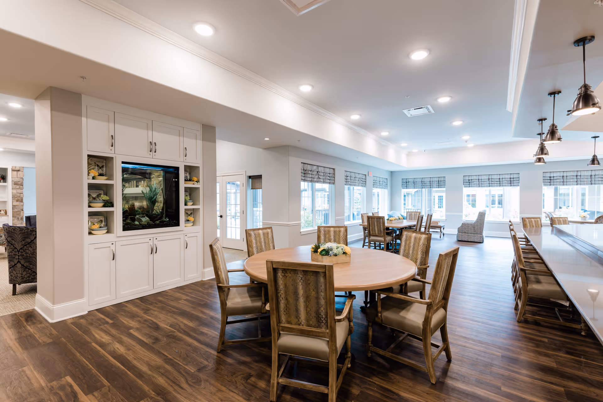A spacious and well-lit common area in a senior living facility featuring a round wooden table with six chairs, additional seating areas with tables and chairs near large windows, and a built-in cabinet with an aquarium. The room has wooden flooring, recessed lighting, and pendant lights over a counter with bar stools.