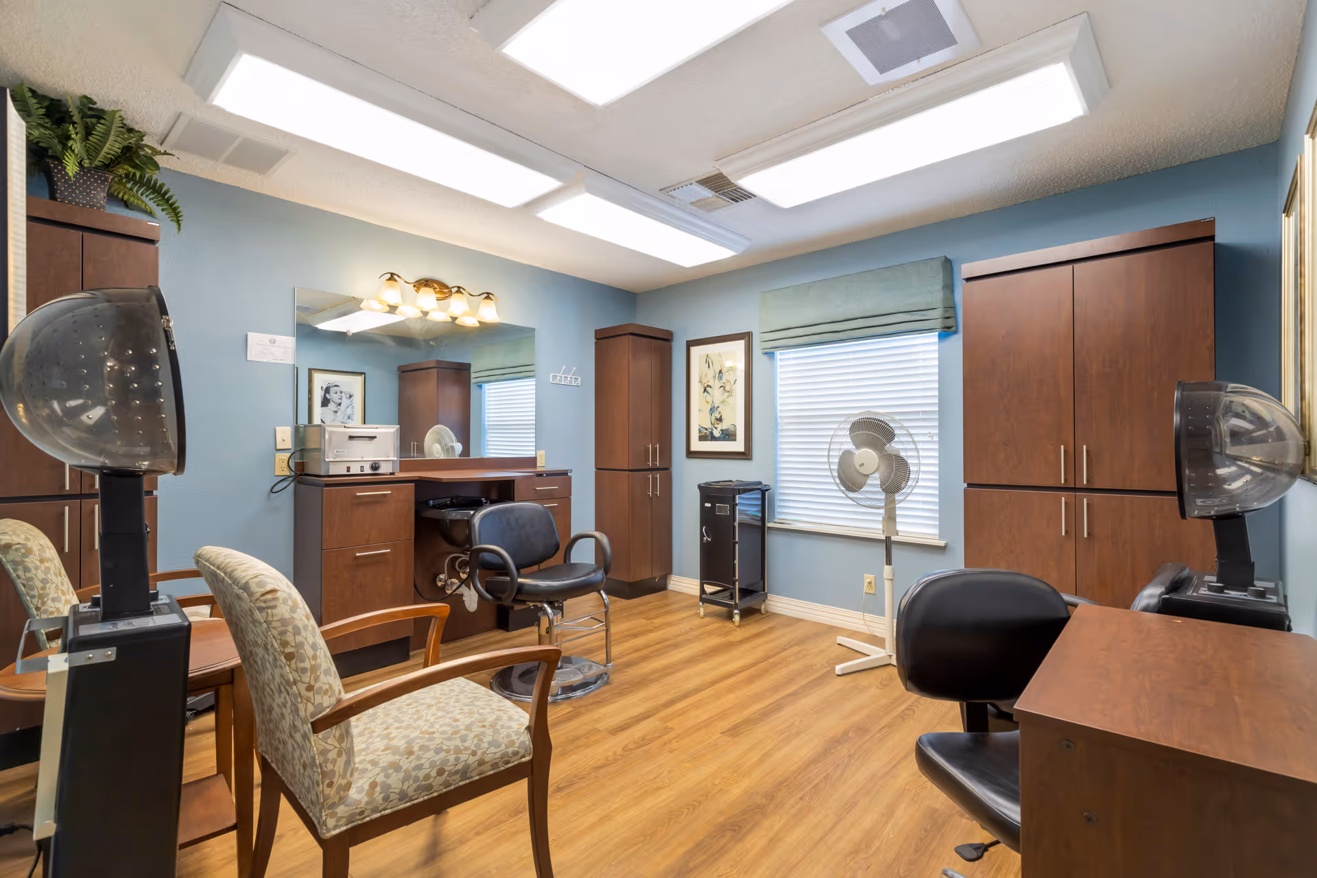 Interior view of a hair salon room with blue walls and wood flooring. The room contains salon chairs, hair dryers, a large mirror with lights above it, wooden cabinets, a standing fan, and framed artwork on the wall.