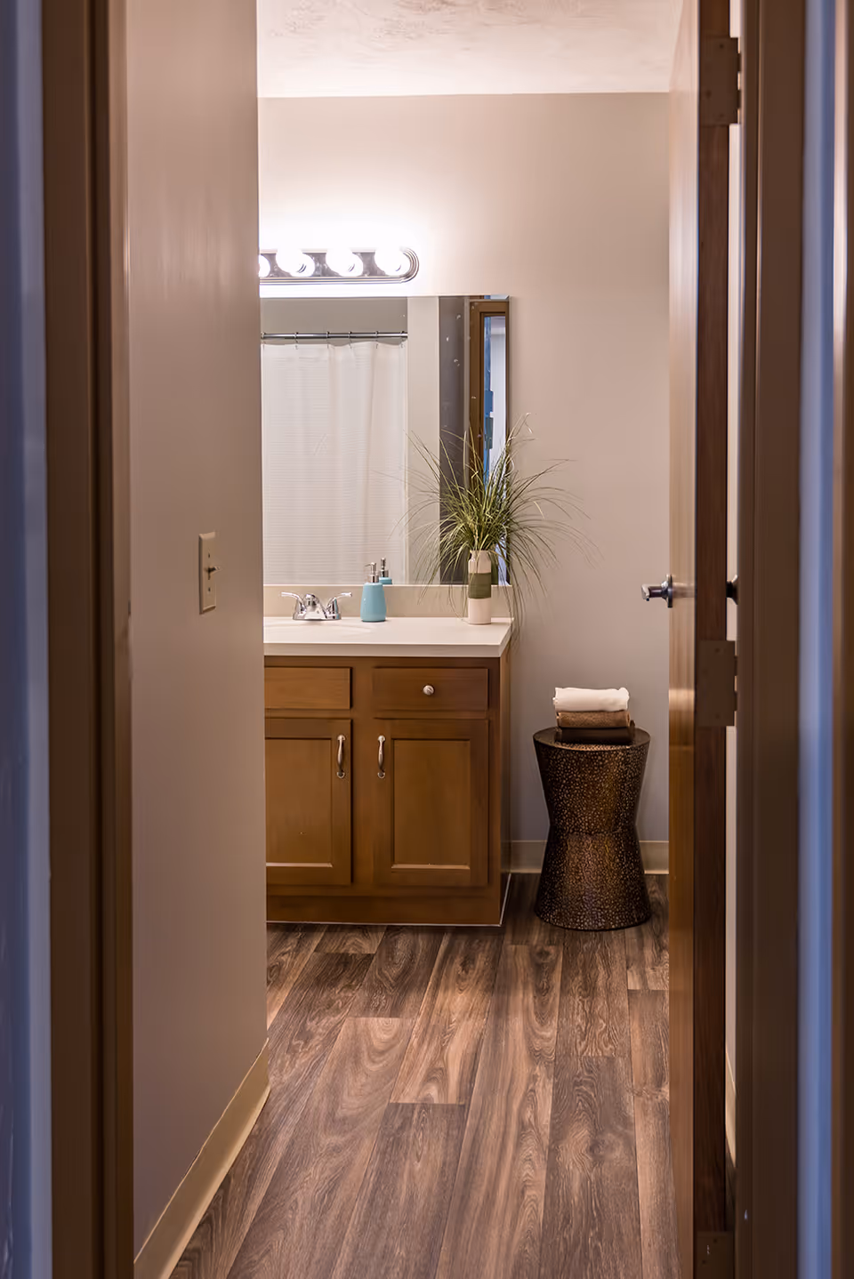 View through a doorway into a bathroom featuring a wooden vanity with a white countertop, a sink with a silver faucet, a soap dispenser, a tall green plant in a white pot, a mirror above the sink, a shower curtain in the background, and a small round table holding folded towels on the right side.