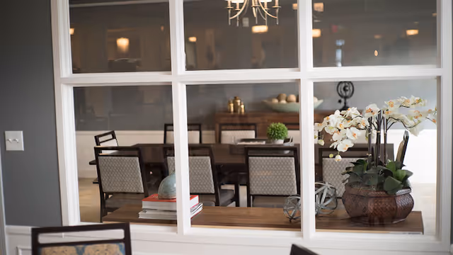 View through a multi-pane interior window into a dining room with a long table, chairs, a chandelier, and decorative plants and books.