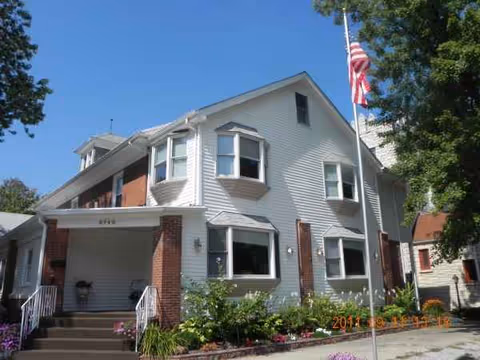 Two-story light-gray building with brick columns, front steps, and an American flag.