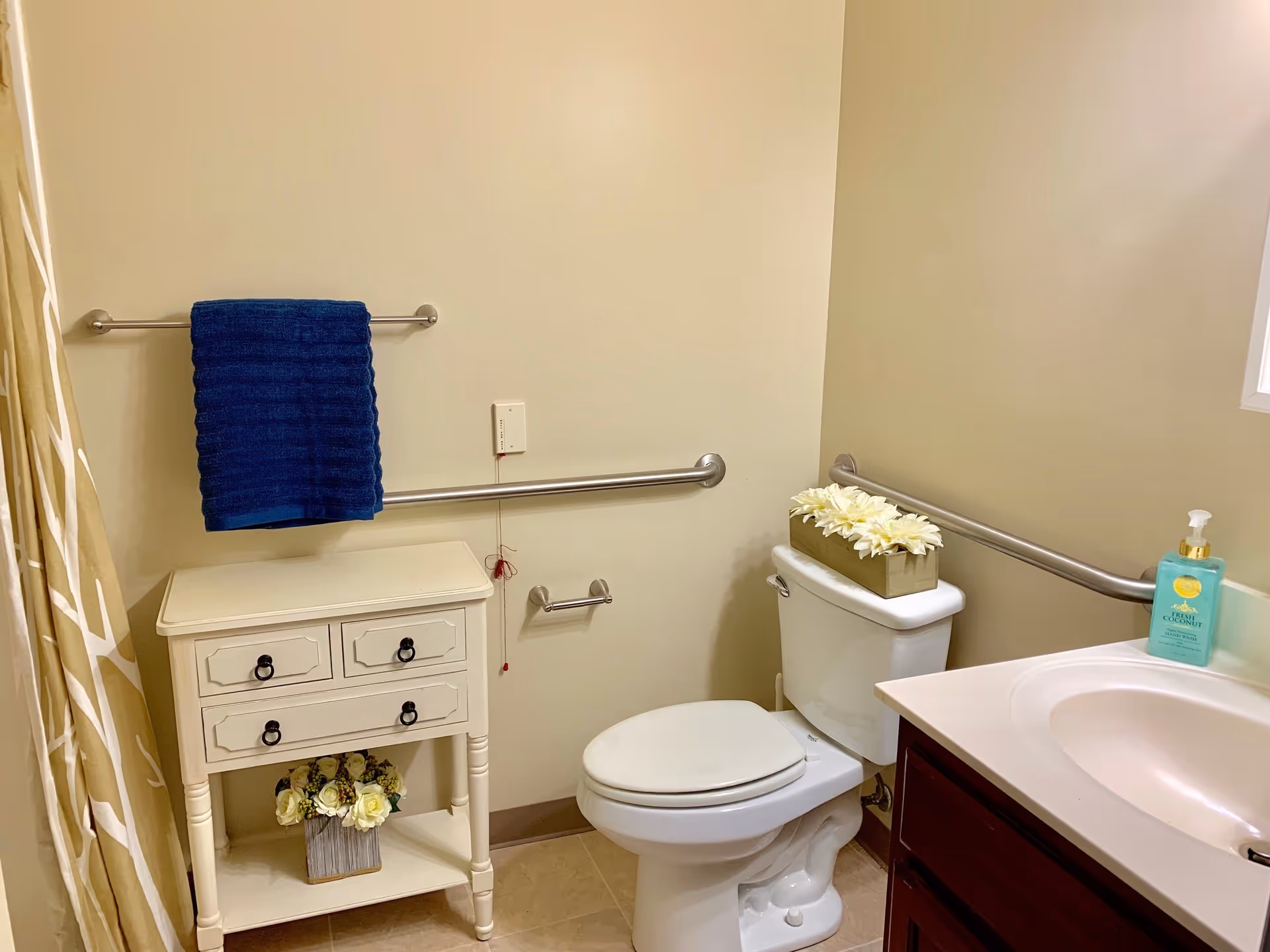 Accessible bathroom with a toilet, sink and vanity, grab bars, a towel on a rack, and a small decorative cabinet with flowers.