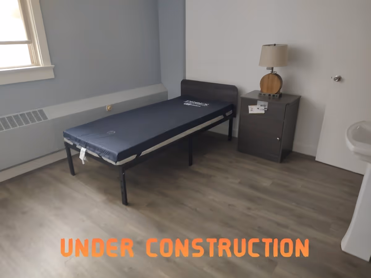 A small, sparsely furnished room with a single bed frame and mattress, a nightstand with a lamp, a window, and a sink. The floor is wood-style laminate, and the walls are painted light gray and white. The words 'UNDER CONSTRUCTION' are overlaid in orange text at the bottom of the image.