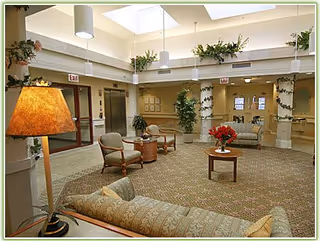 A spacious and well-lit common area in a senior living facility with comfortable seating including armchairs and a sofa, a round wooden table with a vase of red flowers, potted plants, and overhead lighting. The area has carpeted flooring and light-colored walls with large windows and doors in the background.