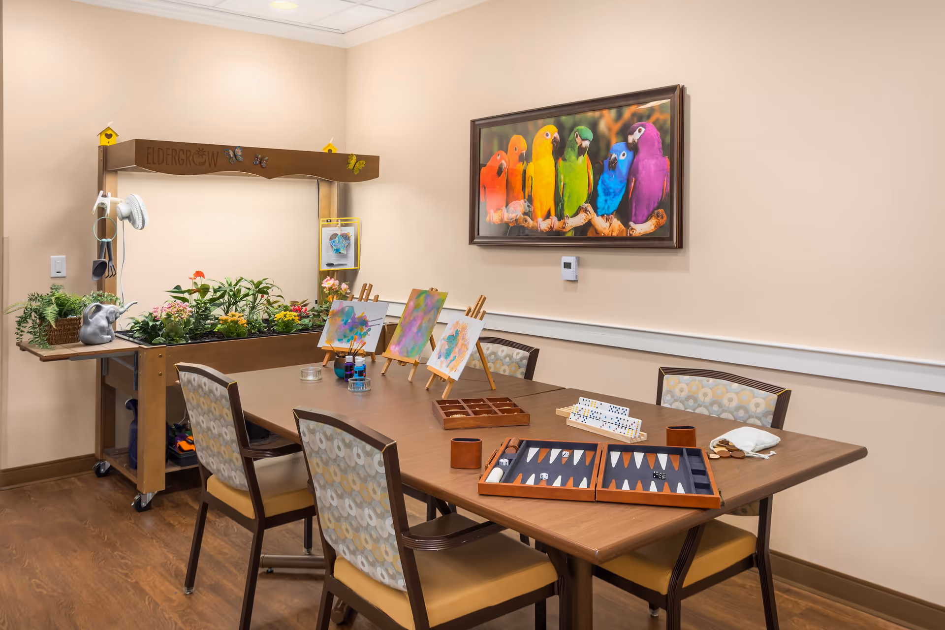 A cozy activity room with a wooden table surrounded by four chairs with patterned upholstery. On the table are three small easels holding colorful paintings, paintbrushes, paint jars, a backgammon set, a domino set, and a small pouch with game pieces. In the corner, there is a gardening station labeled 'Eldergrow' with various plants and flowers. A vibrant framed picture of six colorful parrots hangs on the beige wall above the table.