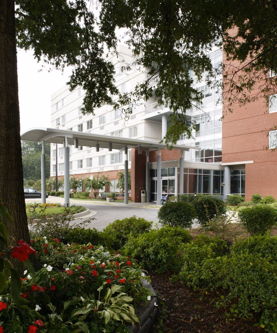 Entrance of a multi-story senior living building with a covered drop-off canopy and landscaped flowerbeds in front.