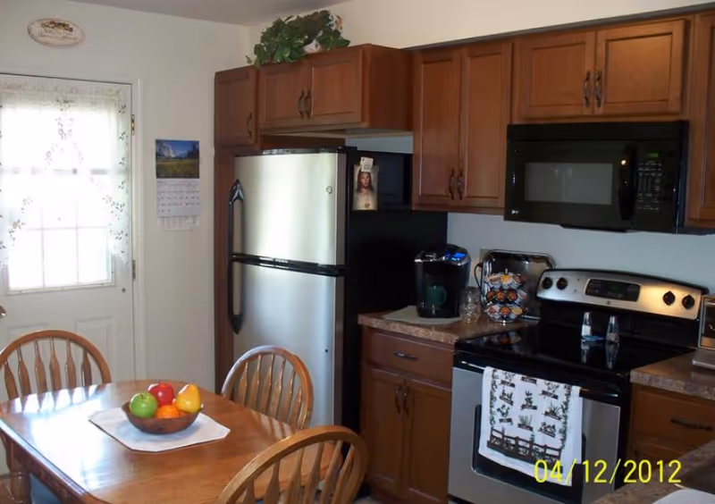 A kitchen with wooden cabinets, a stainless steel refrigerator, a black microwave above a stove, and a coffee maker on the counter. A wooden dining table with four chairs is in the foreground, with a bowl of fruit on a white placemat. A door with a window covered by a lace curtain is visible in the background.