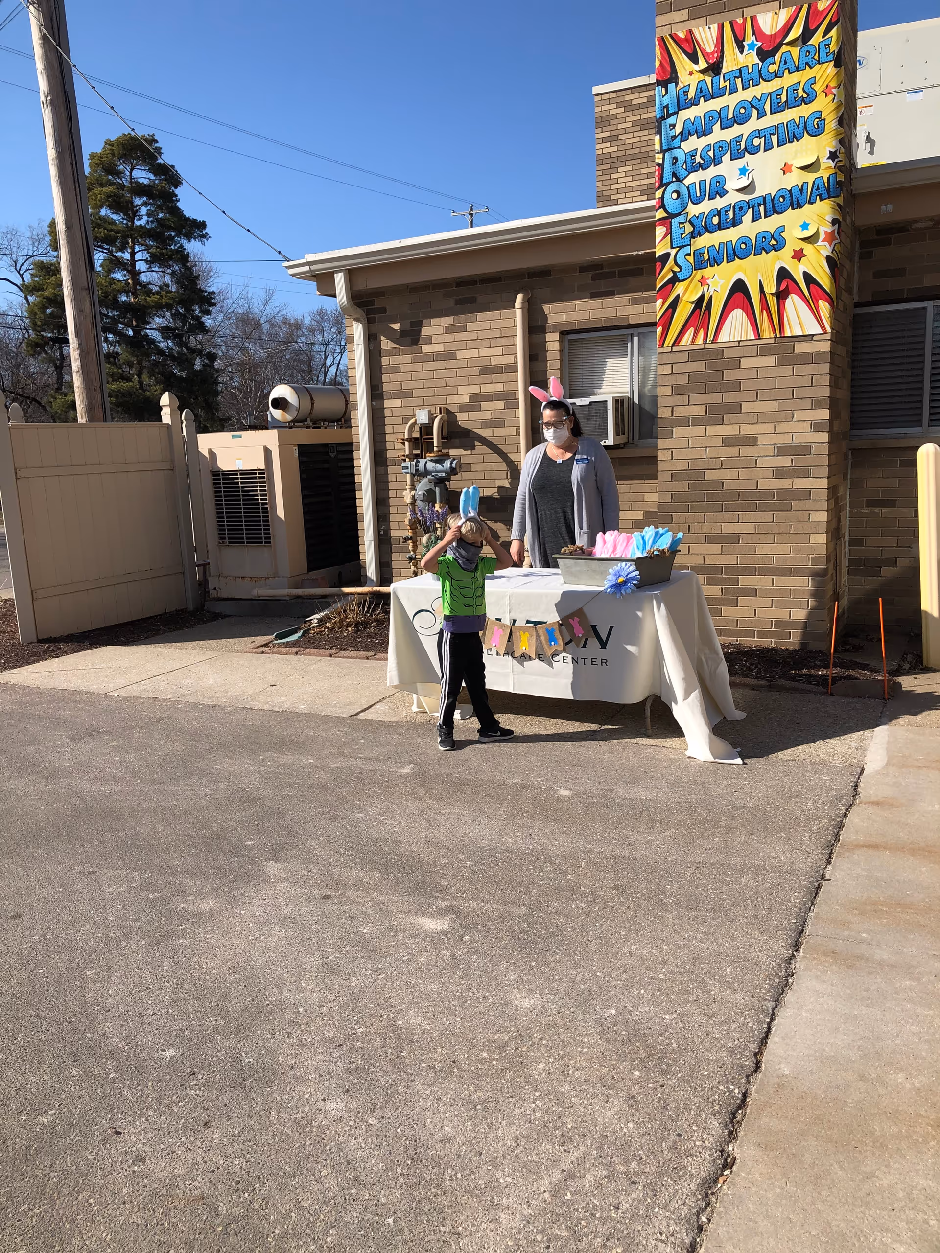 A woman wearing bunny ears and a child stand behind a decorated table outside a brick healthcare building with a colorful banner on the wall.