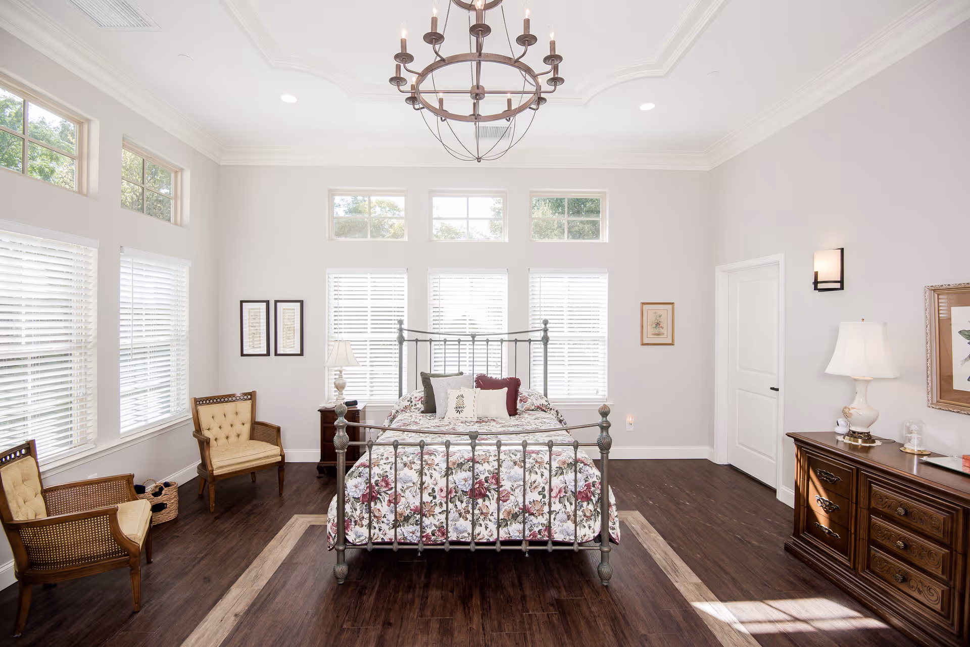 Spacious, sunlit bedroom featuring a metal-framed bed with floral bedding, a chandelier, seating area, and wooden dresser.