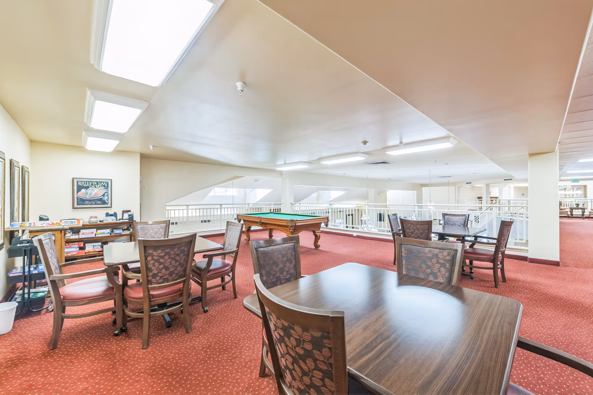 A spacious common area with multiple tables and chairs arranged for social or recreational activities. There is a pool table in the center, a shelf with board games, and bright overhead lighting. The room has red carpet flooring and white walls, with large windows allowing natural light to enter.