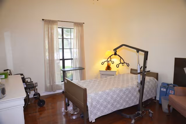 A furnished senior bedroom with a hospital-style bed, patient lift, nightstand with lamp, wheelchair and a window with curtains.