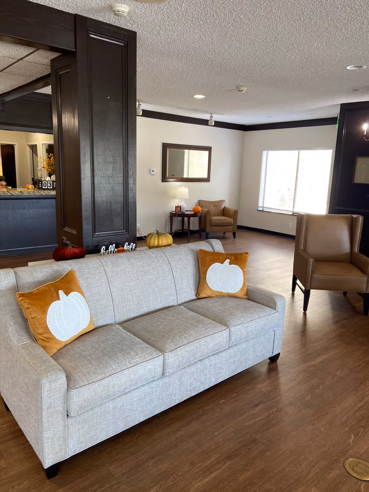A senior living facility common area with a light gray sofa adorned with two orange pillows featuring white pumpkin designs. There are two brown armchairs, a small side table with a lamp and a pumpkin, and a large window letting in natural light. The floor is wooden, and the walls are light-colored with dark trim. A dark wooden column partially separates the seating area from a reception desk in the background.