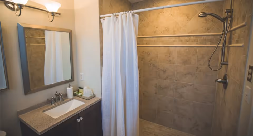 A bathroom with a granite countertop sink, a rectangular mirror above the sink, a wall-mounted light fixture, and a tiled walk-in shower with a white shower curtain and a handheld showerhead.