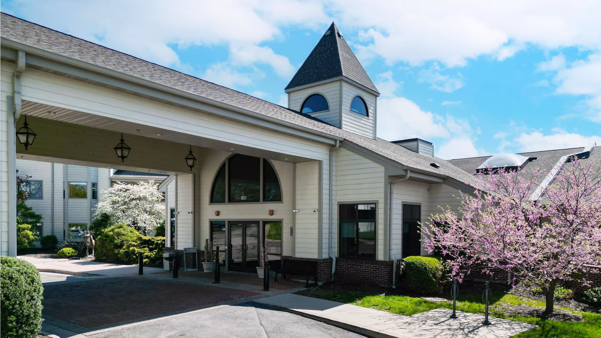 Exterior view of a senior living facility building with a covered entrance, large windows, and a small tower-like structure on the roof. There are blooming trees and well-maintained landscaping around the building under a partly cloudy blue sky.