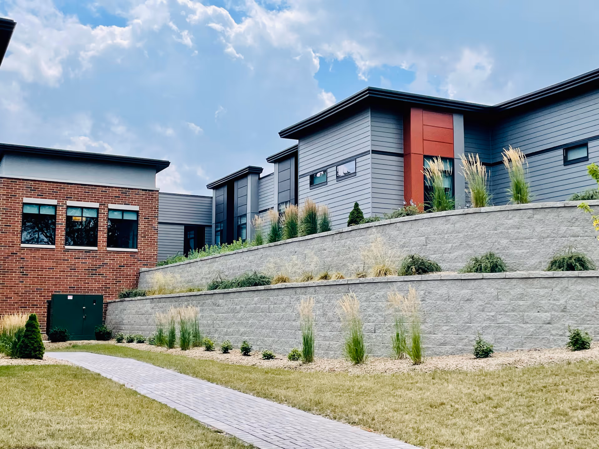 Modern multi-level senior living building with brick and gray siding, terraced stone retaining walls, landscaped lawn and a paved walkway under a partly cloudy sky.