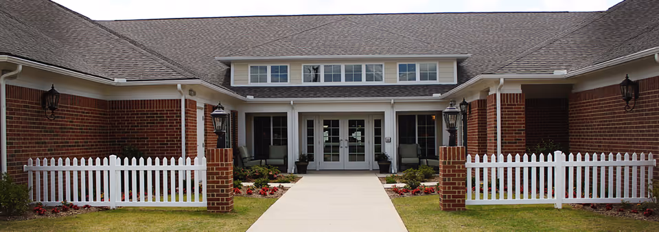 Front exterior view of a senior living facility with red brick walls, white trim, a gray shingled roof, white picket fences on either side of a concrete walkway leading to double glass doors, and outdoor seating areas with chairs near the entrance.