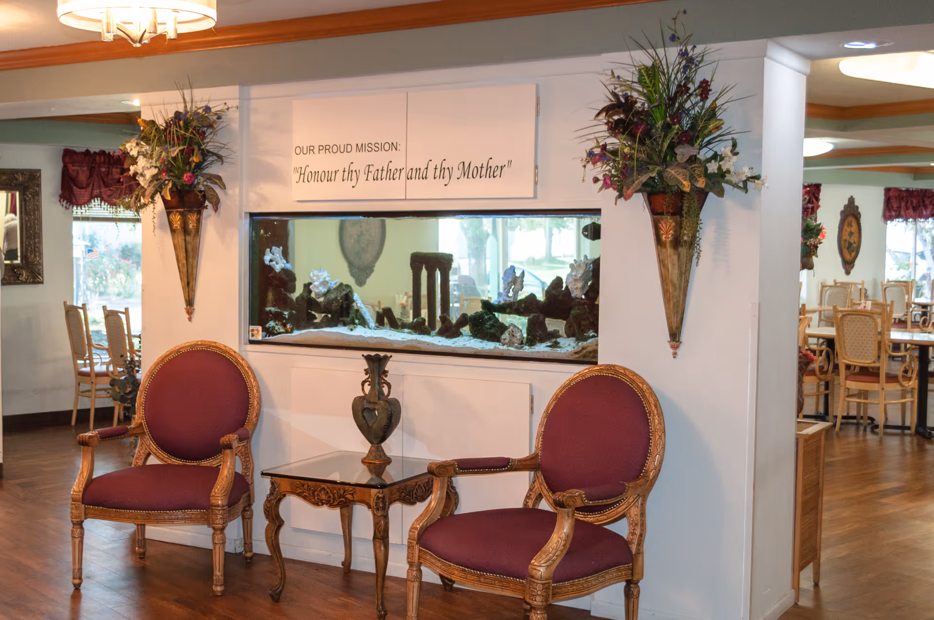 Interior seating area with two ornate wooden chairs with purple cushions and a small glass-top table with a decorative vase. Behind the chairs is a large built-in aquarium with decorative elements inside. Above the aquarium is a sign that reads 'OUR PROUD MISSION: Honour thy Father and thy Mother'. The walls have floral arrangements in wall-mounted vases, and in the background, there is a dining area with tables and chairs.