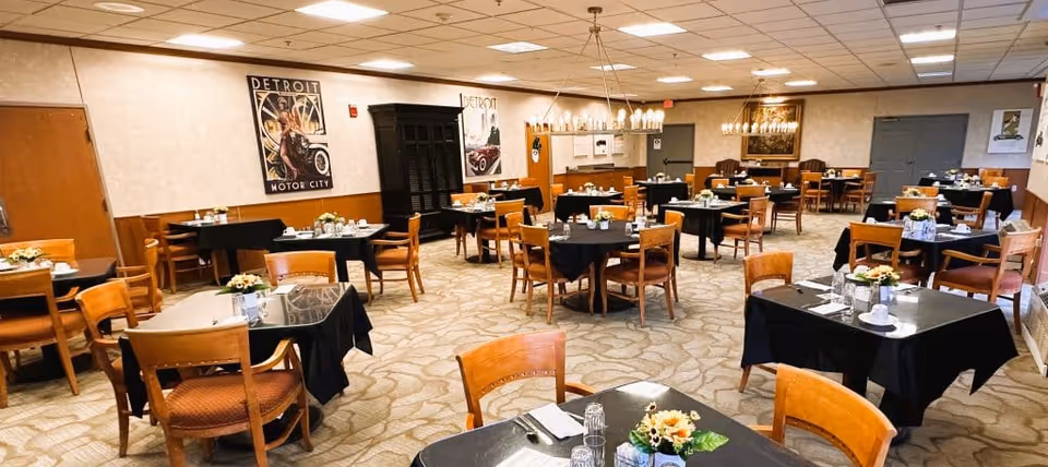 A spacious dining room with multiple tables covered in black tablecloths, each set with glassware, napkins, and small floral centerpieces. Wooden chairs surround the tables. The room has beige walls with vintage Detroit Motor City posters, a carpeted floor, and ceiling lights providing bright illumination.