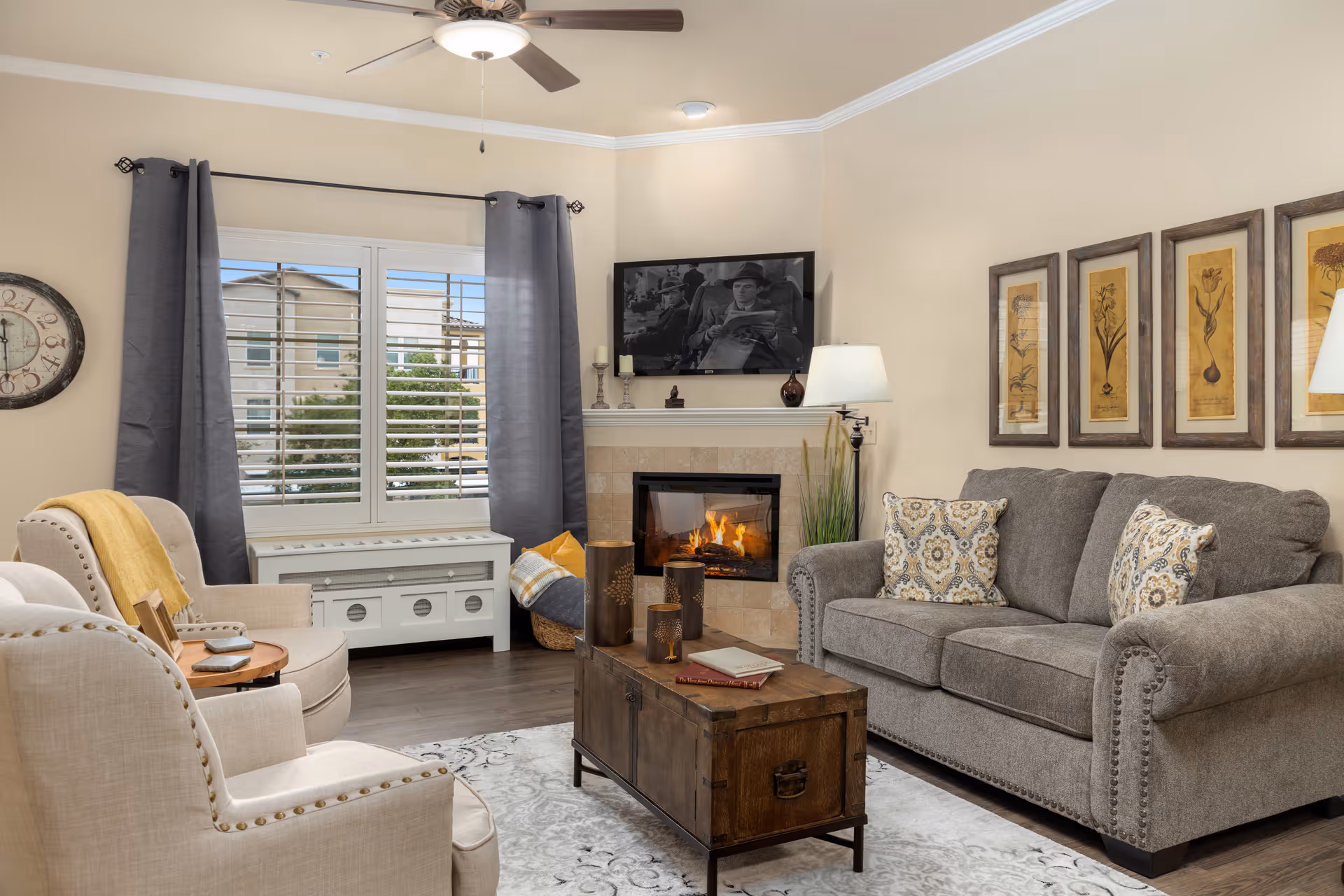 A cozy living room with a gray sofa adorned with patterned pillows, two beige armchairs with nailhead trim, a wooden coffee table with decorative items and books, a fireplace with a fire burning, a wall-mounted TV above the fireplace, a window with gray curtains and white shutters, a wall clock, and framed botanical artwork on the wall.