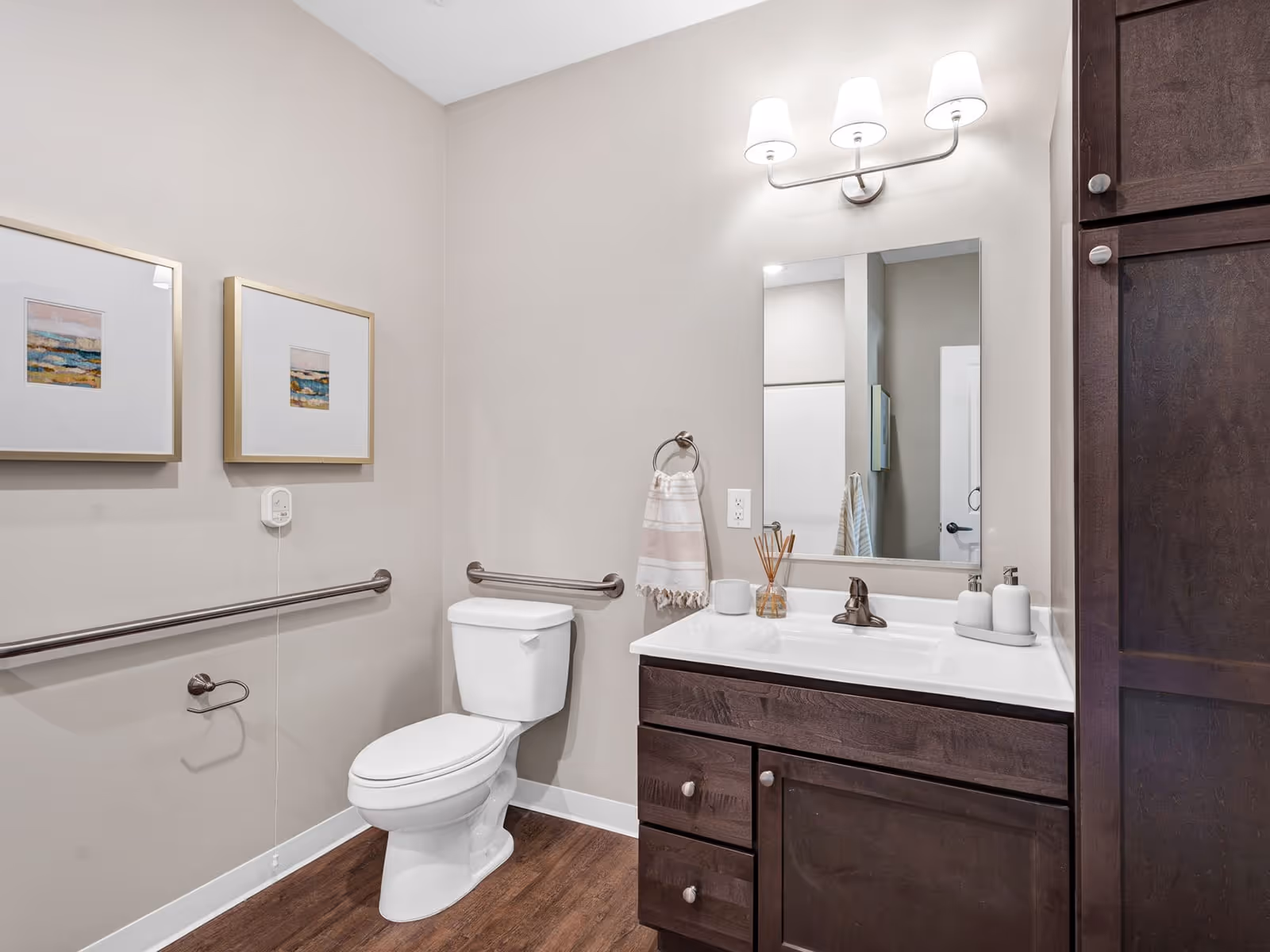 A clean and modern bathroom with a white toilet, dark wood vanity with a white countertop, a rectangular mirror above the sink, and a three-light wall fixture. The walls are painted light beige and decorated with two framed abstract paintings. There are metal grab bars installed on the walls near the toilet for accessibility, and a towel ring with a striped hand towel next to the sink. The floor has a wood-like finish.