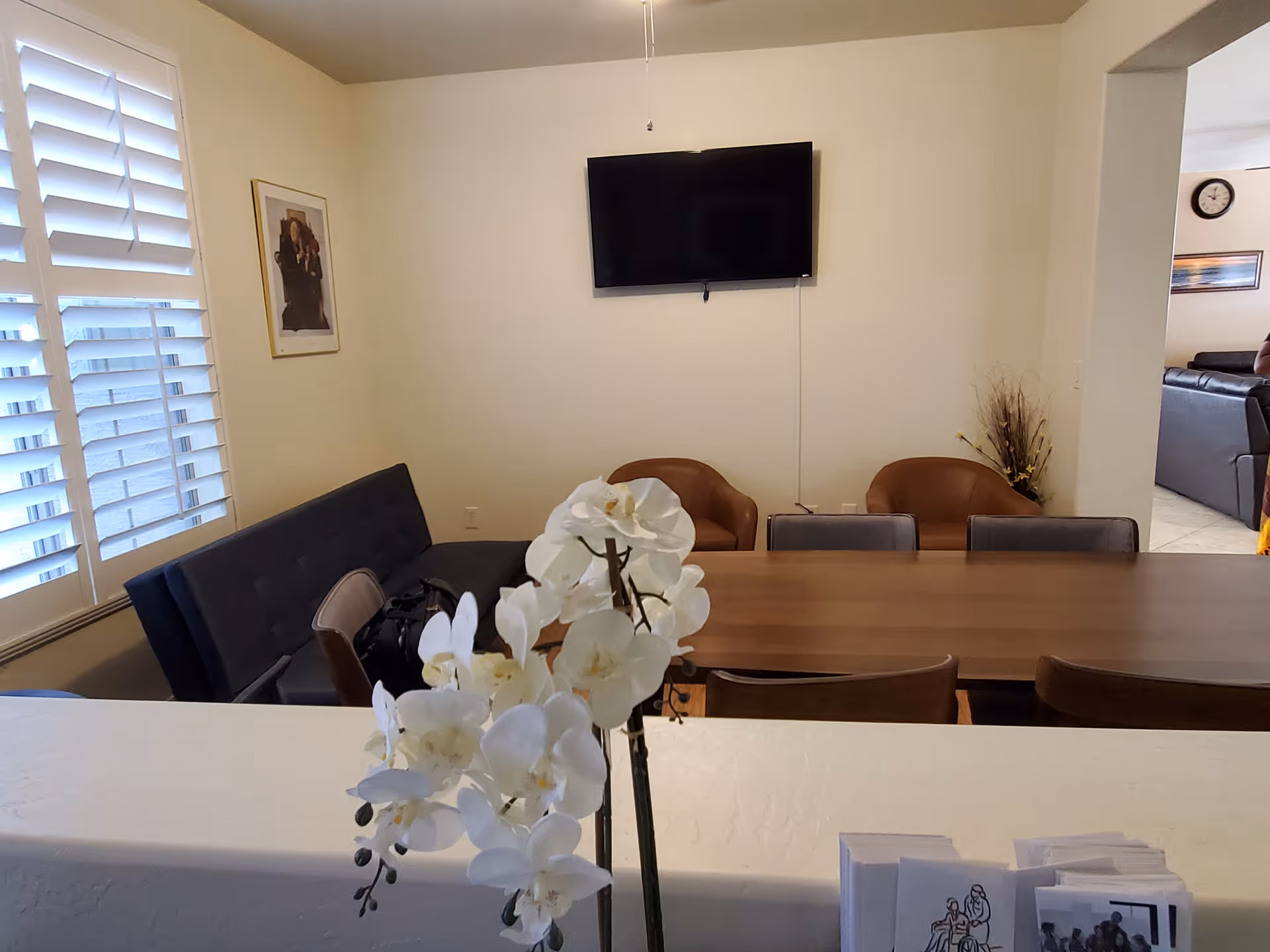 Interior view of a room with a wooden dining table surrounded by chairs and two brown armchairs against the wall. A flat-screen TV is mounted on the wall above the armchairs. A window with white shutters is on the left side, and a framed picture hangs on the wall. In the foreground, there is a white orchid flower arrangement on a counter with some brochures or pamphlets.