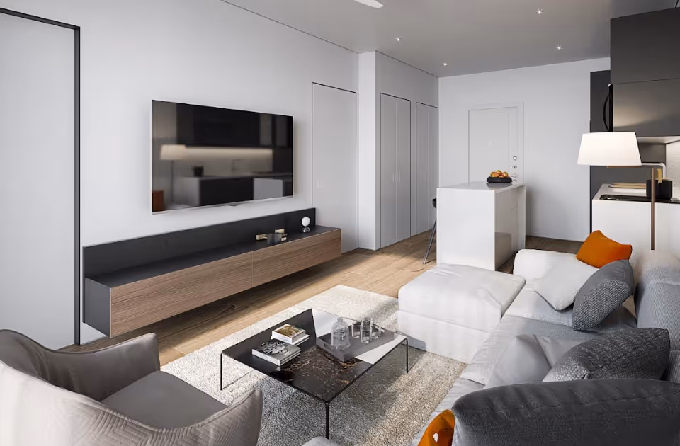 Modern living room with a large gray sectional sofa adorned with gray and orange pillows, a glass coffee table with books and glassware, a wall-mounted flat-screen TV above a wooden and black media console, and a small kitchen area with a white island and bar stools in the background.