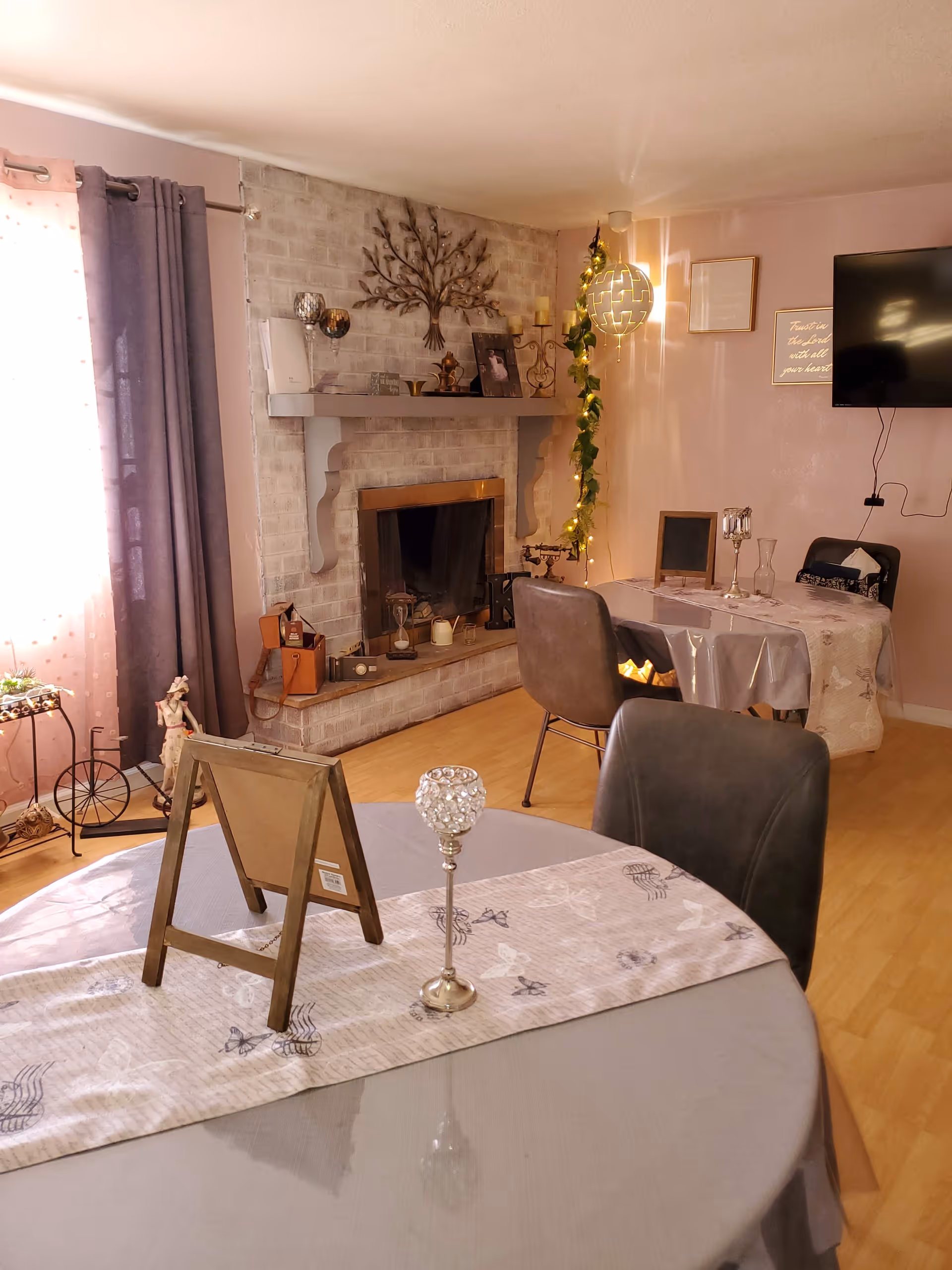 A cozy living room and dining area with a light-colored brick fireplace decorated with a metal tree wall art and various small decorative items. Two tables with gray tablecloths and patterned runners are set with candle holders and picture frames. There are gray chairs around the tables, a wall-mounted TV, and soft lighting including a hanging light fixture with greenery. A window with sheer and dark curtains lets in natural light.