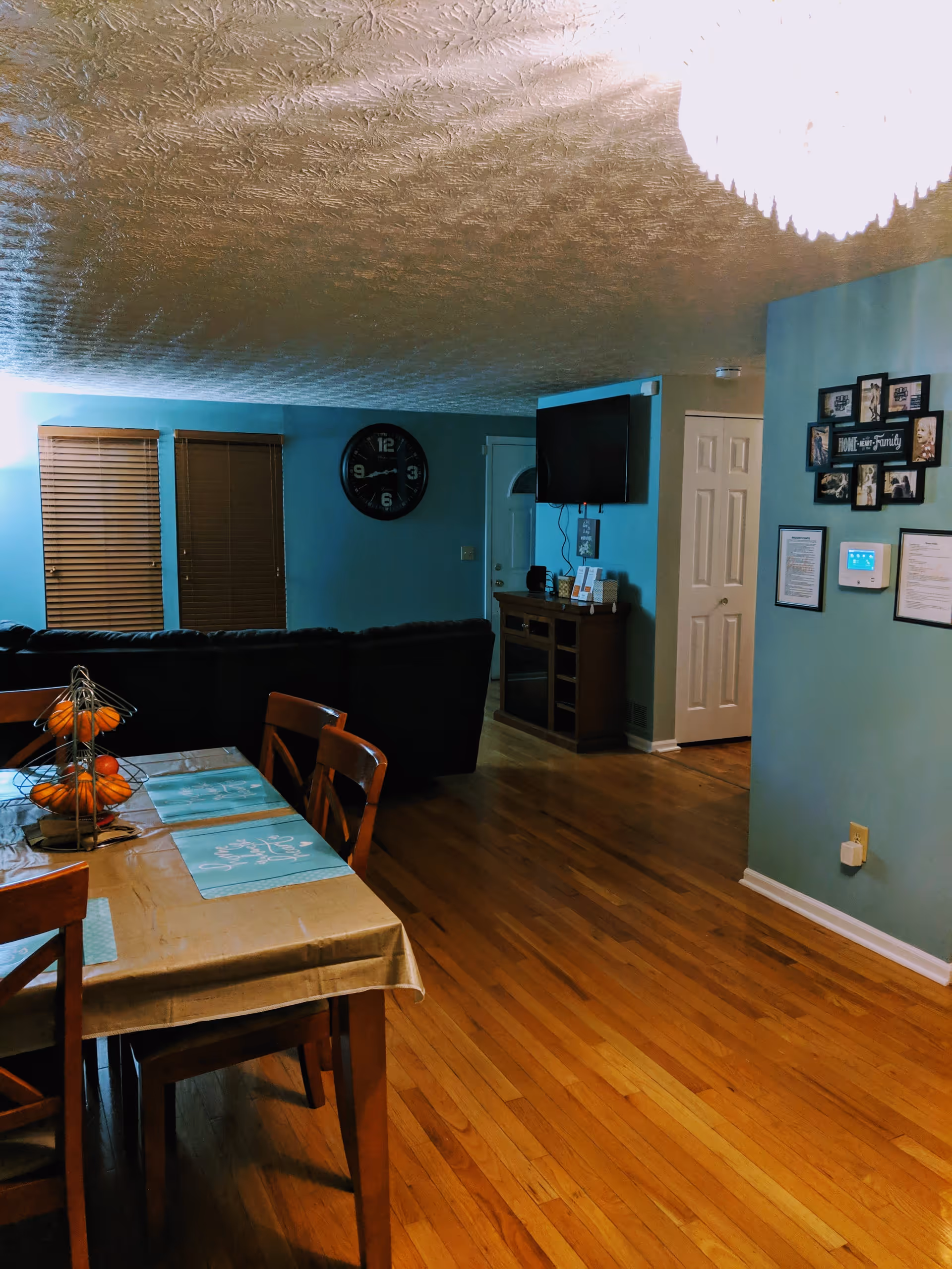 Interior view of a senior care home living and dining area with wooden floors, a dining table with chairs and placemats, a black couch, a wall-mounted TV, a clock on the wall, and family photos displayed on a blue wall.