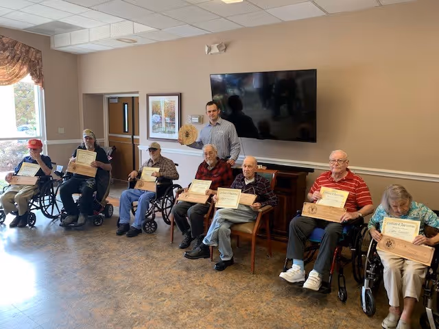 A group of elderly individuals sitting in a room, most in wheelchairs, holding certificates of achievement. A younger man stands behind them holding a wooden plaque. The room has beige walls, a large flat-screen TV mounted on the wall, and a window with curtains letting in natural light.