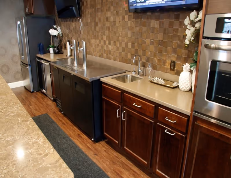 A modern kitchen area with wooden cabinets, a stainless steel refrigerator, built-in oven, two sinks, and a countertop with a decorative tray and white pineapple ornament. The backsplash features a textured, square-patterned design, and there is a mounted TV on the wall.