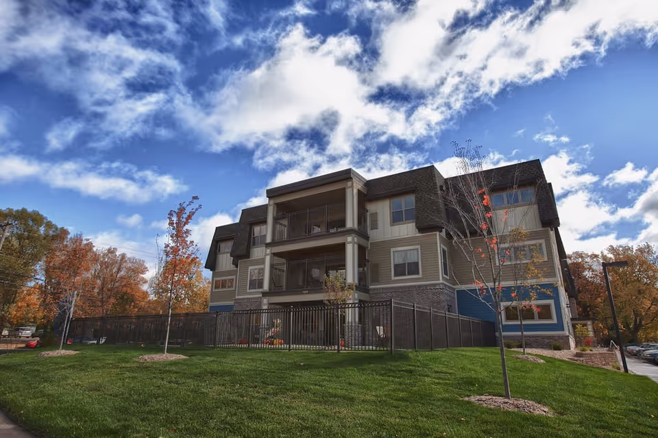 Exterior view of a three-story assisted living and memory care facility building with balconies, surrounded by a black metal fence, green grass, and young trees with autumn foliage under a partly cloudy blue sky.