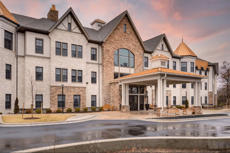 Exterior view of a large senior living facility building with a covered entrance, multiple windows, and a combination of stone and light-colored siding. The sky is at sunset with pink and orange hues, and there are benches and small landscaped areas near the entrance.