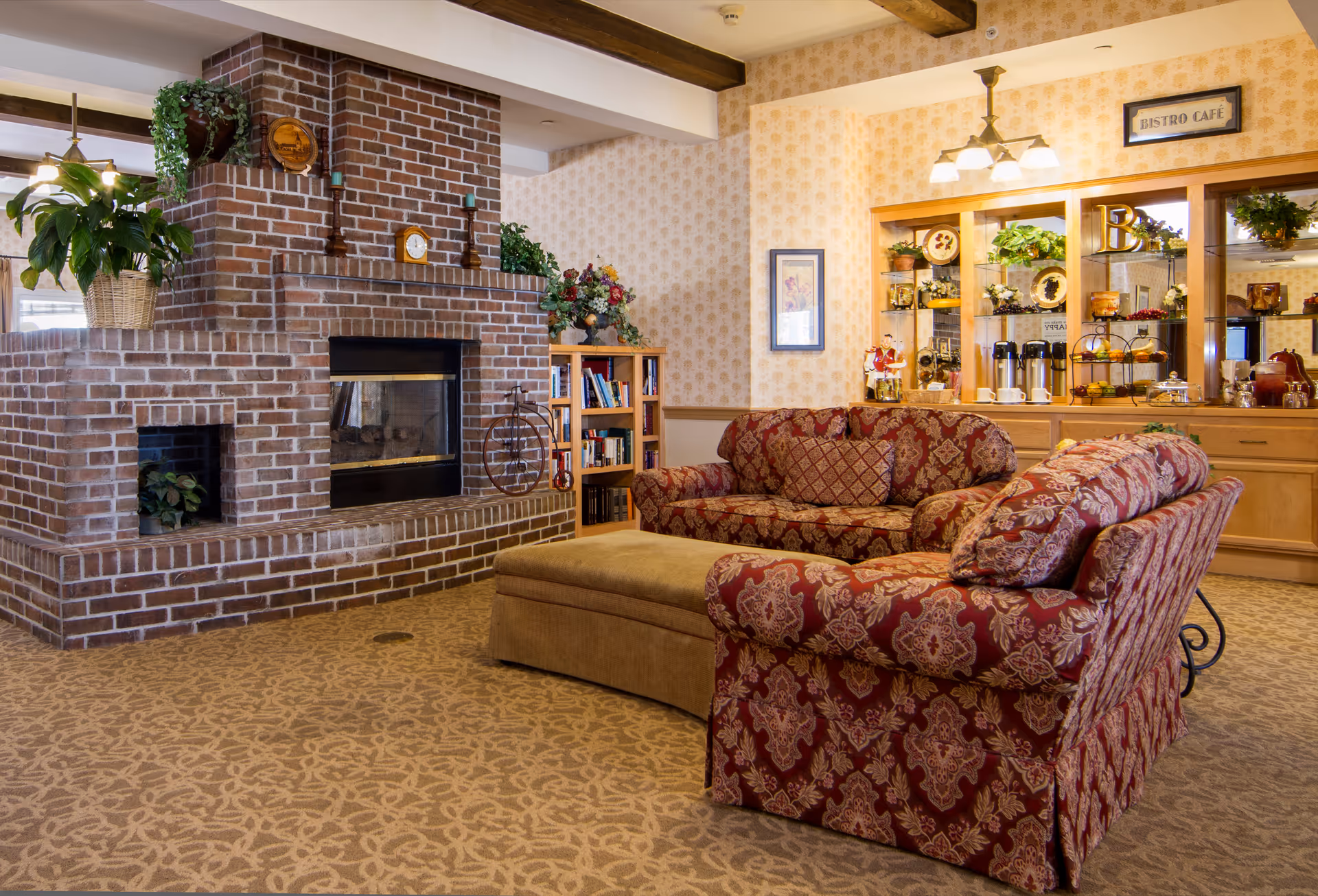 A cozy living room area with patterned red sofas and a beige ottoman in front of a large brick fireplace. The room features a bookshelf with books and decorative plants, a wall with framed artwork, and a wooden cabinet labeled 'Bistro Café' displaying coffee pots, cups, and decorative items. The carpet has a floral pattern and the ceiling has wooden beams.