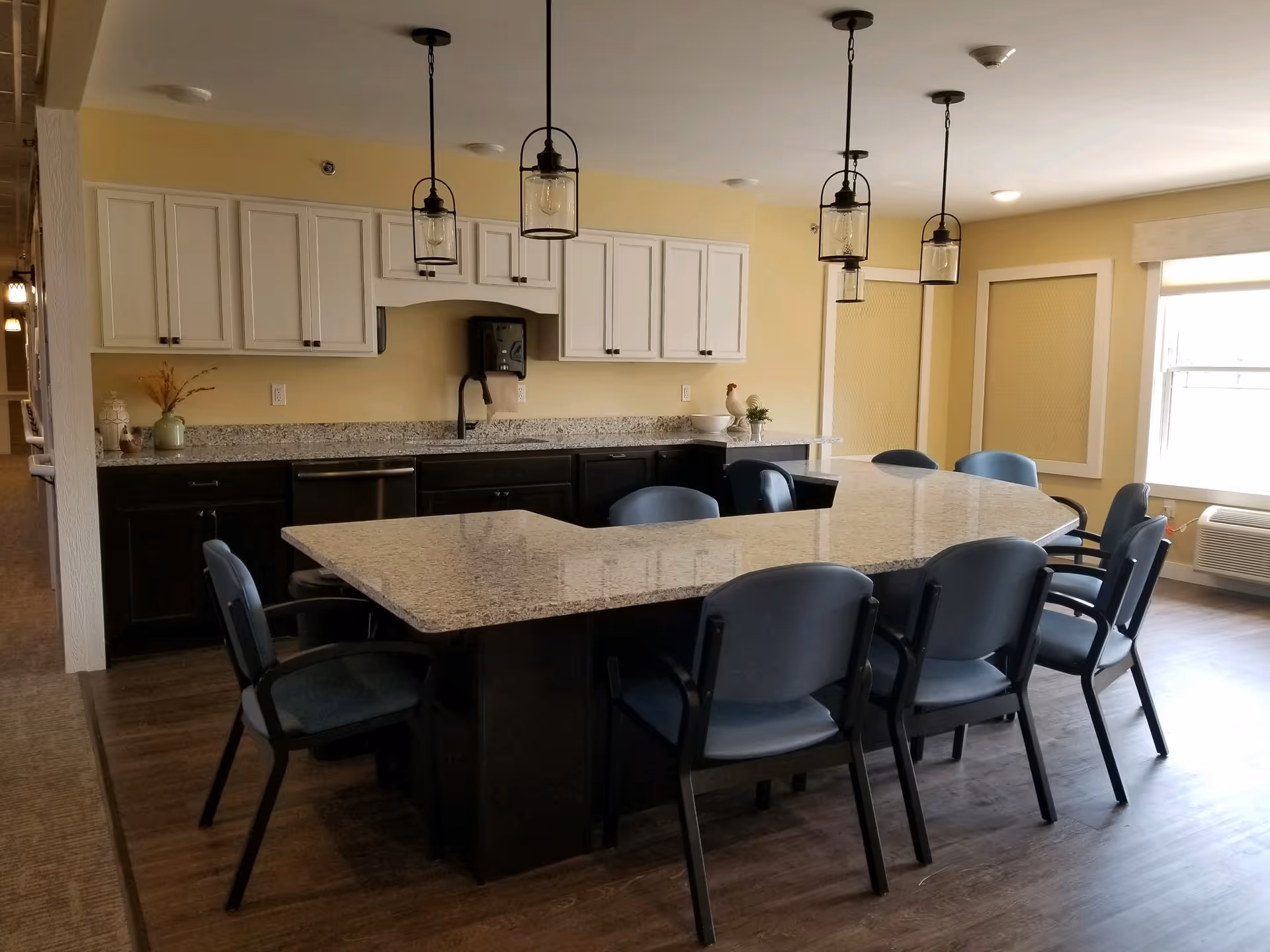 A modern kitchen and dining area with a large granite island surrounded by eight blue chairs. The kitchen features white upper cabinets, dark lower cabinets, a stainless steel dishwasher, and three pendant lights hanging above the island. There is a window on the right side letting in natural light.