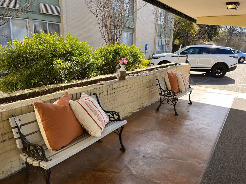 Outdoor covered seating area with two white benches featuring black metal armrests and legs, each bench adorned with large orange and white striped cushions. A low brick wall with a vase of pink flowers separates the seating area from green bushes. In the background, a white SUV is parked near a building with windows and air conditioning units.